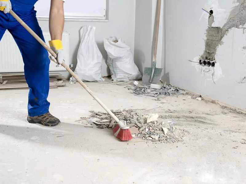 A worker sweeping rubble and plaster dust from the floor of a stripped-out room